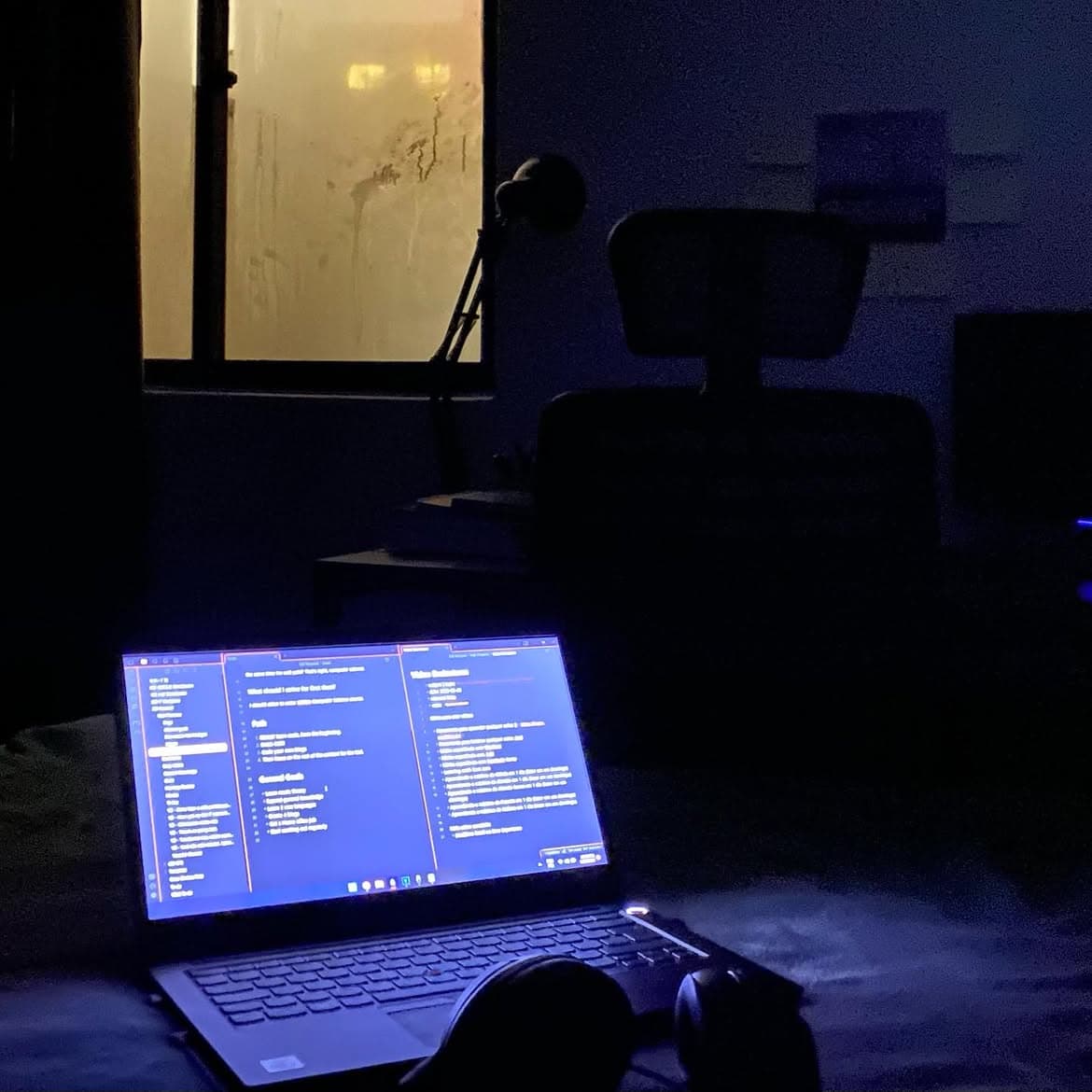 Quarto escuro com um notebook aberto sobre a cama, a tela azul iluminando o ambiente, e a janela amarelada ao fundo