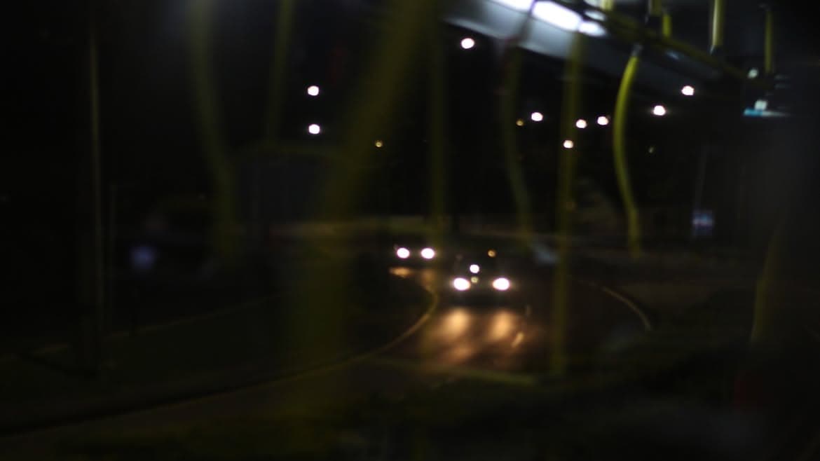Foto desfocada feita de dentro de um ônibus à noite: faróis de carros numa curva, com as barras amarelas em primeiro plano