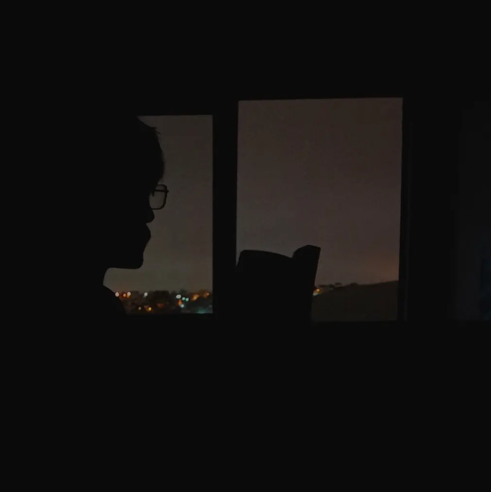 Silhueta de uma pessoa de óculos diante da janela de um quarto escuro, com as luzes da cidade lá fora